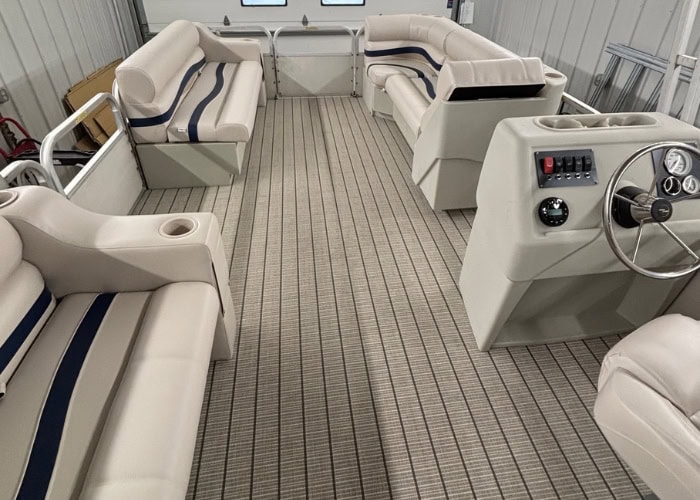 Interior of a boat with cream-colored cushioned seating, striped flooring, and a helm with a steering wheel and controls on the right.