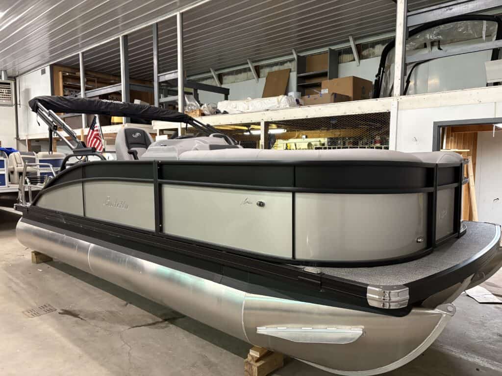 A sleek 2025 Barletta Aria pontoon boat with black and gray panels is parked indoors, showcasing plush seating and an American flag at the rear.