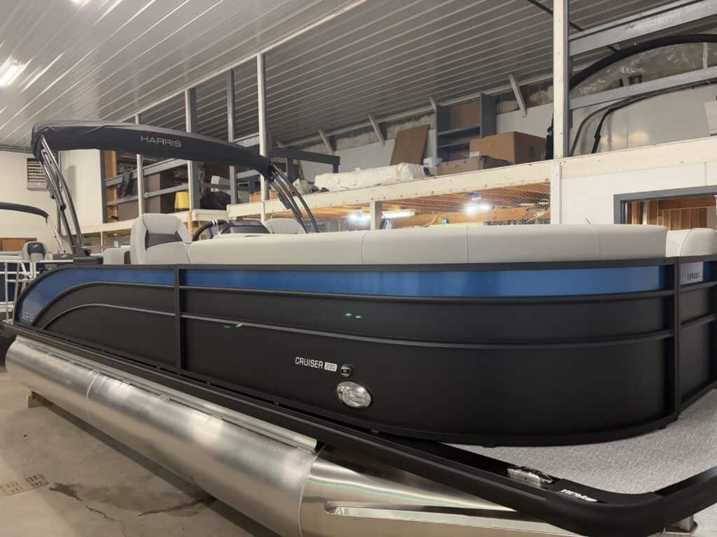 The 2025 Harris Cruiser 230 SLDH, a sleek blue and black pontoon boat with comfortable gray seating, is parked indoors under a sturdy metal roof structure.