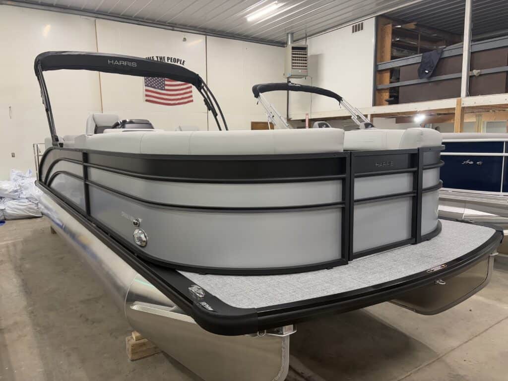 A Harris Cruiser 230 pontoon boat with gray panels and black trim is parked in a garage, where an American flag hangs proudly on the wall in the background.