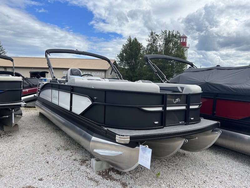 The 2024 Barletta Cabrio 24UE, a sleek black and white pontoon boat, is displayed outdoors on gravel. An overcast sky looms overhead with trees visible in the background.