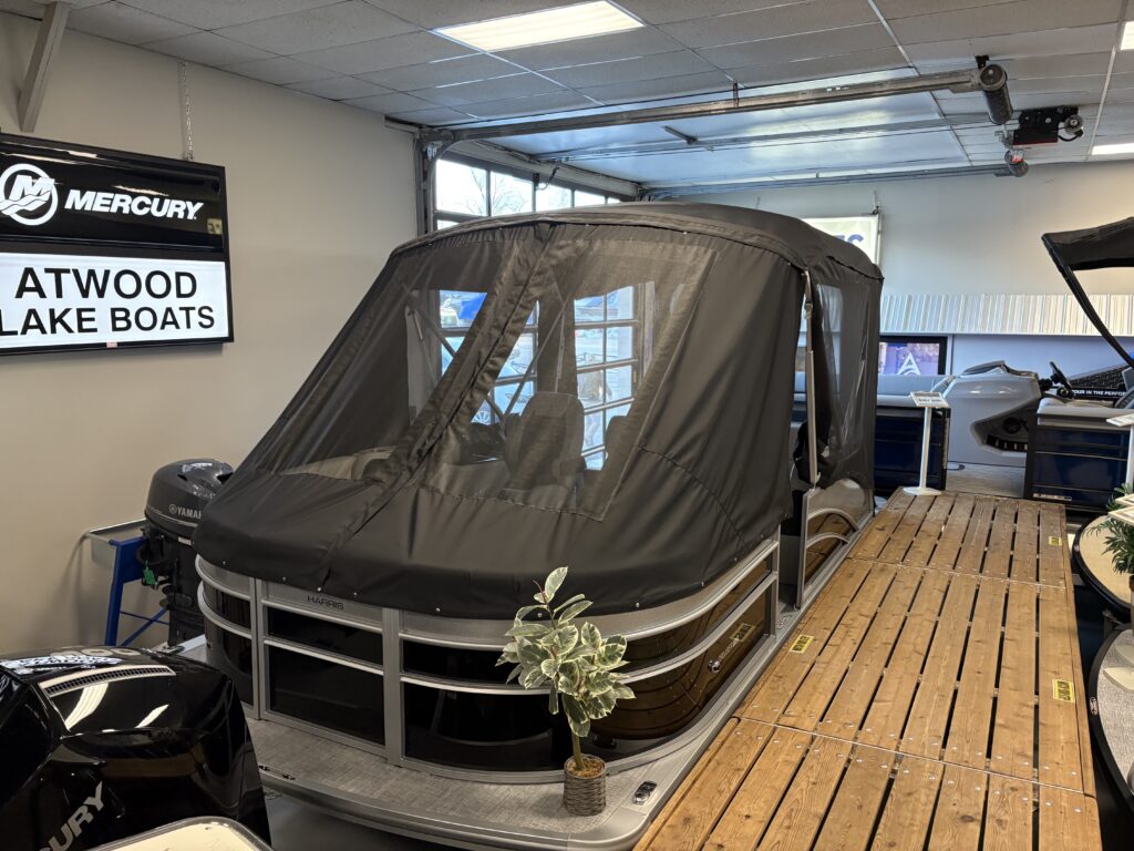 A sleek Harris Cruiser pontoon boat with a black canopy cover is parked inside a garage. A wooden walkway runs beside it, framed by a small plant in the foreground, creating an inviting and serene setting.