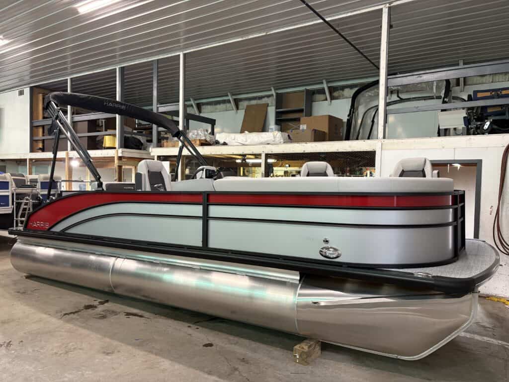 The 2025 Harris Cruiser 230, with its red and gray exterior, is parked indoors. This pontoon boat boasts cushioned seats and a sturdy canopy frame, promising both style and comfort for your next adventure on the water.