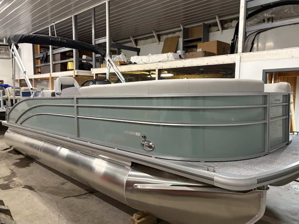 A light blue and gray Harris Cruiser pontoon boat is parked indoors. There are boxes and other boats, including a 230 CS, visible in the background, hinting at a busy 2025 boating season ahead.